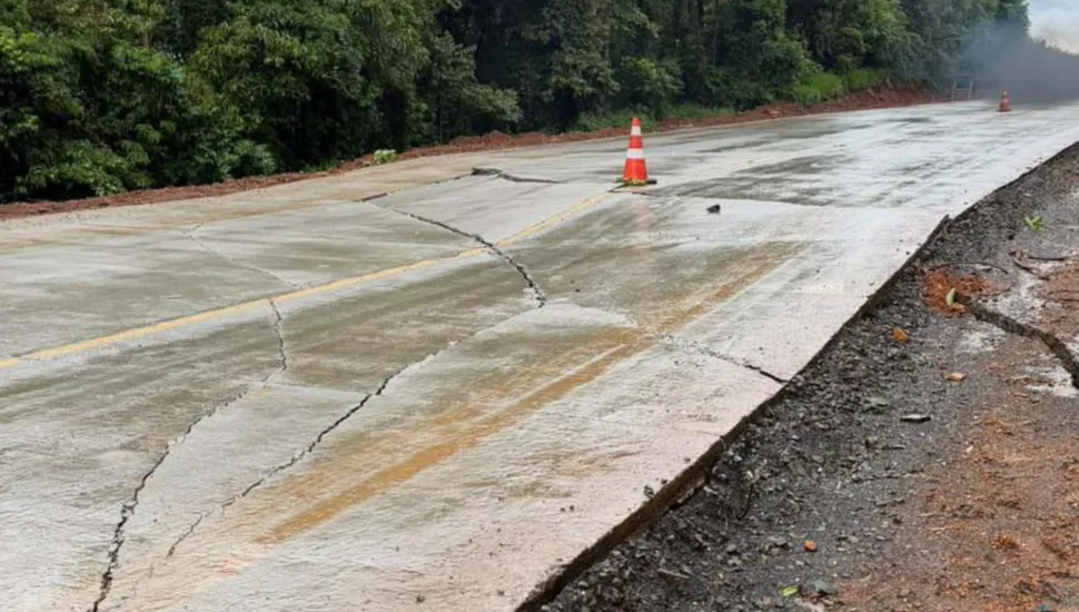 Fotografia em plano médio de uma rodovia pavimentada em concreto que apresenta rachaduras e danos estruturais na pista. Em primeiro plano, a laje de concreto da estrada exibe grandes fissuras transversais e longitudinais que atravessam a faixa de rolamento, sob uma superfície molhada. Um cone de sinalização laranja e branco encontra-se posicionado sobre a pista danificada para alertar motoristas. No plano intermediário, a rodovia estende-se em curva suave, ladeada por encostas de terra vermelha e vegetação densa. Ao fundo, sob um dia nublado e chuvoso, árvores e arbustos compõem a paisagem marginal da estrada afetada.