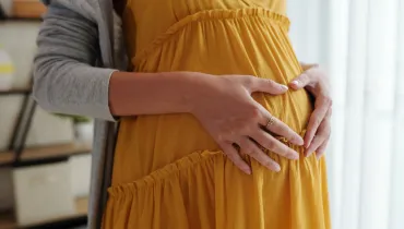 Fotografia em primeiro plano (close-up) de uma gestante acariciando a barriga, captada em um ambiente interno iluminado por luz natural difusa. Em primeiro plano, as mãos de uma mulher repousam delicadamente sobre sua barriga proeminente, formando o contorno de um coração com os dedos unidos. Ela veste um vestido amarelo de tecido leve com detalhes franzidos e camadas, além de um cardigã cinza sobre os ombros. Um anel dourado é visível em seu dedo anelar. No plano intermediário, o torso da gestante ocupa a porção central da composição, destacando a textura do vestuário e o gesto afetuoso. Ao fundo, uma cortina branca translúcida e elementos borrados de um cômodo residencial ou consultório compõem a iluminação suave da cena.