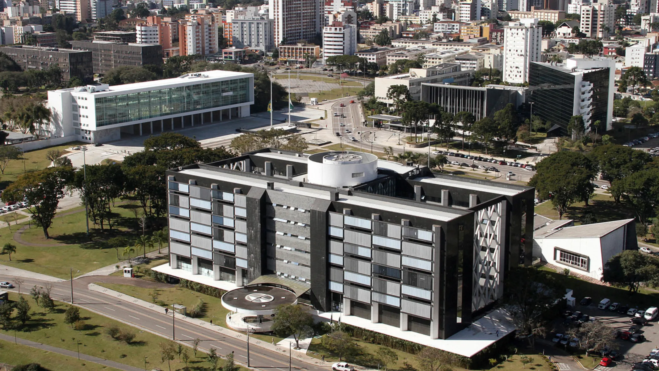 Fotografia aérea em plano geral do Centro Cívico em Curitiba, destacando o edifício da Assembleia Legislativa do Paraná (ALEP) em primeiro plano e o Palácio Iguaçu ao fundo. Em primeiro plano, o imponente edifício da ALEP exibe sua arquitetura moderna com revestimento escuro, janelas alinhadas e uma estrutura circular característica na cobertura e na entrada principal, cercado por áreas verdes arborizadas e vias pavimentadas. No plano intermediário, a esplanada conecta o complexo legislativo ao Palácio Iguaçu, o edifício governamental de fachada branca e extensa estrutura envidraçada, ladeado por praças e estacionamentos. Ao fundo, a densa paisagem urbana de Curitiba estende-se com diversos prédios residenciais e comerciais verticais sob luz solar diurna.