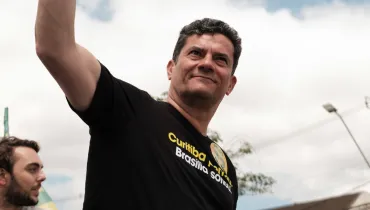 Fotografia em primeiro plano (close-up) captada de baixo para cima do senador paranaense Sergio Moro durante uma manifestação ou evento público ao ar livre. Em primeiro plano, Sergio Moro, um homem de pele clara e cabelos escuros, sorri levemente e olha para cima, com o braço direito erguido em aceno para a multidão (fora de quadro)[cite: 16]. Ele veste uma camiseta preta de algodão com estampas em letras amarelas e brancas, onde se lê claramente "Curitiba rima..." e, abaixo, "...Brasília sonhou", além de um pequeno brasão ou distintivo amarelo no peito. No canto inferior esquerdo, parte do rosto de outro homem mais jovem com barba é visível. No plano intermediário, o ombro e o braço estendido do político ocupam parte do enquadramento, com a textura do tecido e a pele em destaque. Ao fundo, a iluminação natural diurna sob um céu parcialmente nublado e cinzento compõe a atmosfera do local, com postes de luz e fiação elétrica borrados compondo o ambiente urbano.