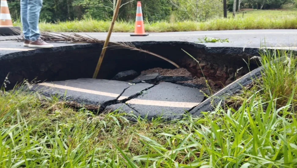 Fotografia em primeiro plano de uma rodovia asfáltica danificada por um colapso estrutural que provocou uma cratera profunda na pista. Em primeiro plano, o acostamento coberto por grama verde estende-se até a borda de um grande buraco na estrada. No plano intermediário, o asfalto cedeu e desabou, revelando uma cavidade subterrânea escura sob a pista; placas de pavimentação com faixas amarelas de sinalização encontram-se fraturadas e caídas no interior da cratera. Na margem superior, cones de sinalização laranjas e brancos estão posicionados ao lado de um pedestre calçando tênis e calça jeans. Ao fundo, a vegetação rasteira e arbustos compõem a paisagem marginal da rodovia sob luz natural diurna.