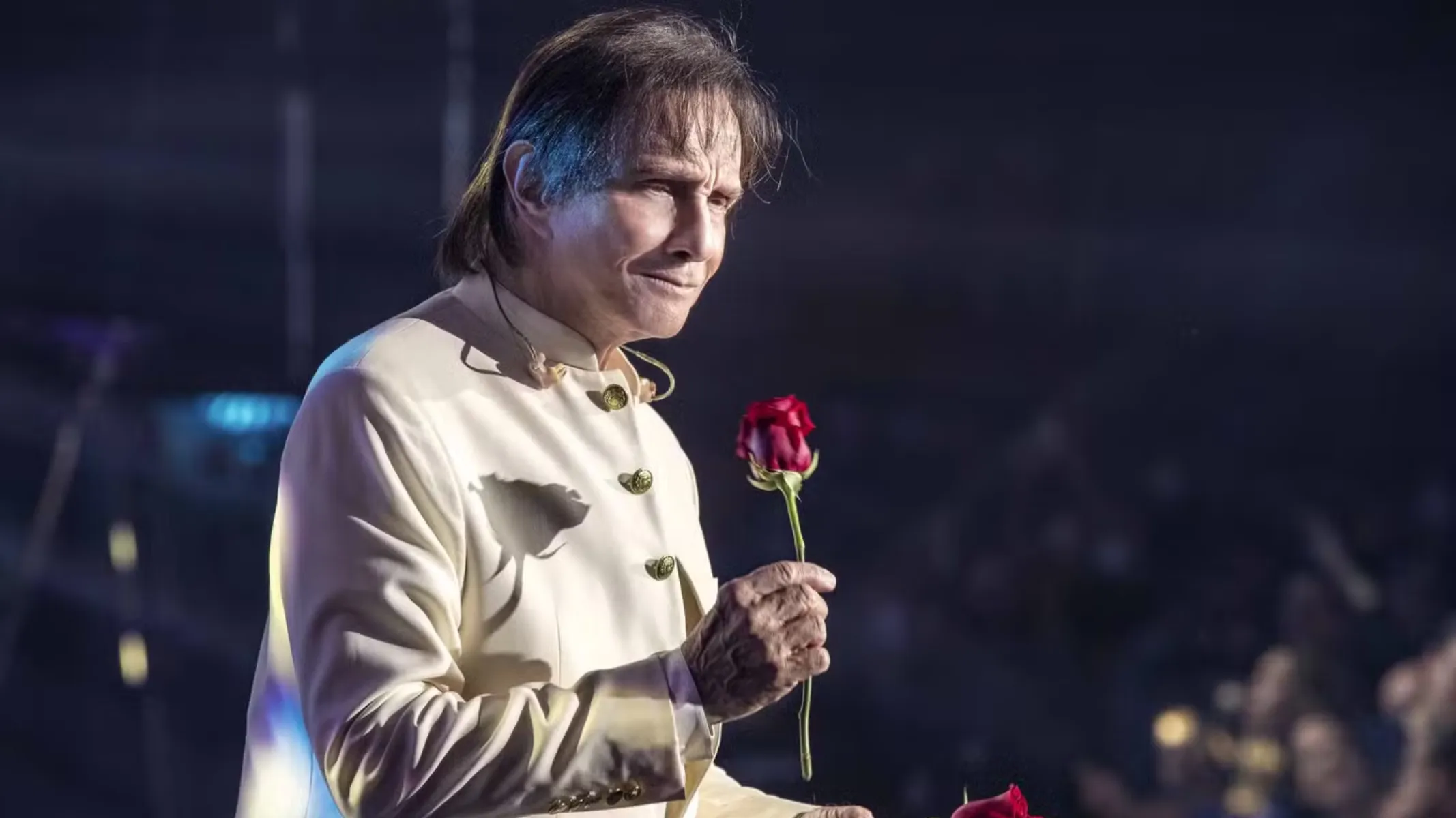 Fotografia em plano médio de Roberto Carlos caracterizado em um show ou apresentação ao ar livre, segurando uma rosa vermelha. Em primeiro plano e ao centro, o cantor brasileiro aparece sorrindo levemente com expressão nostálgica, vestindo sua tradicional camisa social clara de gola alta com botões frontais decorados. Ele segura delicadamente uma rosa vermelha de haste longa com a mão direita, enquanto uma segunda rosa aparece parcialmente no canto inferior. No plano intermediário, a iluminação cênica destaca a figura do artista, projetando uma sombra suave de flor sobre o tecido de sua vestimenta. Ao fundo, a plateia e o ambiente do show permanecem em penumbra, com pontos de luz difusos sob a atmosfera noturna do evento.