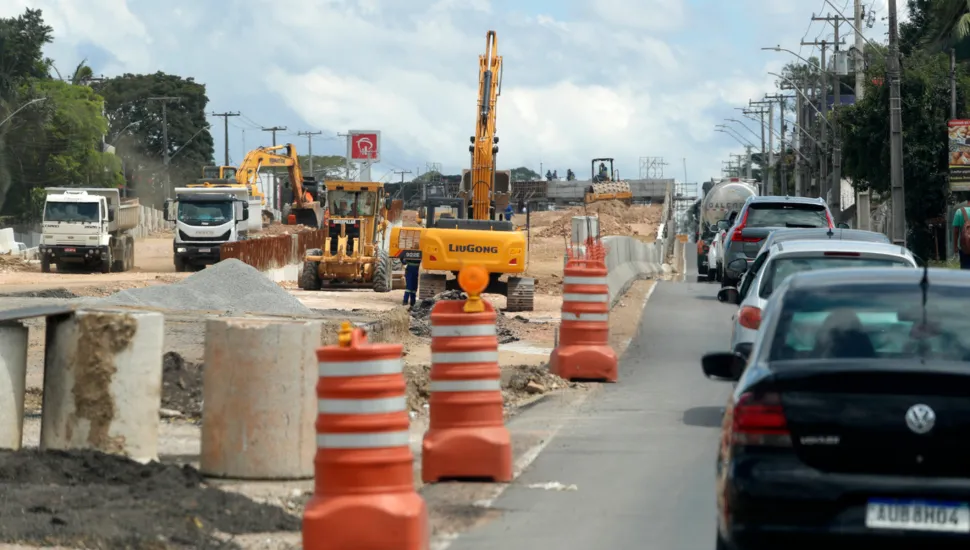 Fotografia em plano geral de obras de infraestrutura e duplicação em uma rodovia ou avenida, com tráfego intenso de veículos retido e maquinário pesado em operação. Em primeiro plano e à esquerda, blocos de concreto e tambores sinalizadores laranjas e brancos delimitam a área de intervenção na pista. À direita, uma longa fila de carros — incluindo um automóvel escuro Volkswagen em destaque — aguarda no fluxo de trânsito lento. No plano intermediário, diversos equipamentos de construção civil pesada atuam no canteiro de obras, destacando-se escavadeiras hidráulicas amarelas (uma delas da marca LiuGong), pás carregadoras, caminhões basculantes e pilhas de brita e terra. Ao fundo, postes de rede elétrica, vegetação lateral e um céu parcialmente nublado completam o cenário de obras viárias sob luz diurna.