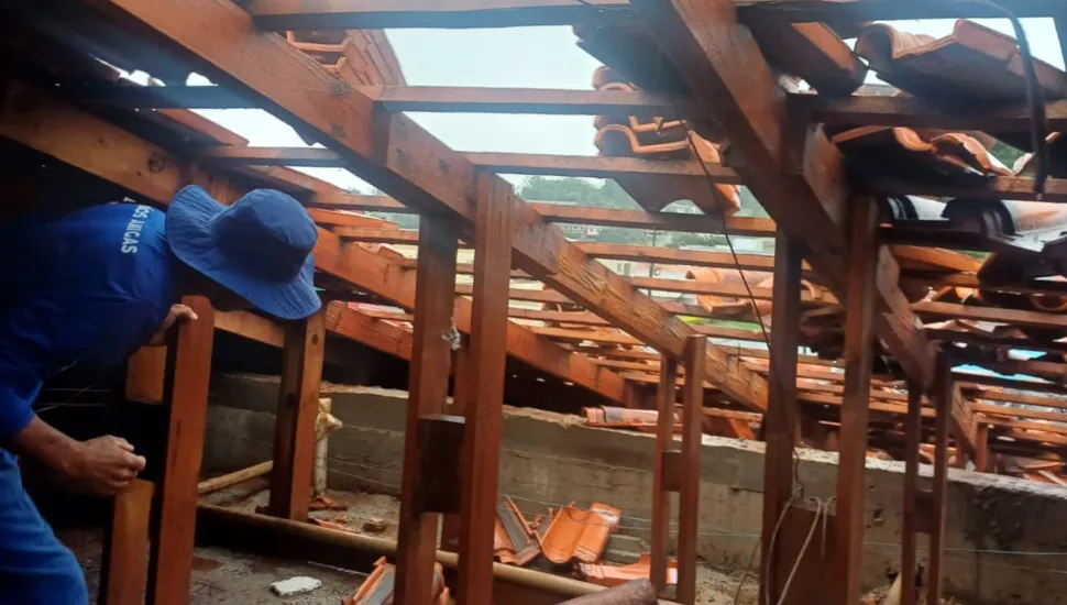 Fotografia em plano médio de um trabalhador inspecionando a estrutura de madeira danificada de um telhado sob o qual há entulhos e telhas quebradas. Em primeiro plano e à esquerda, um trabalhador vestindo uniforme azul e chapéu de abas largas azul apóia-se em uma viga de sustentação de madeira, avaliando os danos na cobertura. No plano intermediário, o telhado exibe vigas, caibros e ripas de madeira expostas, com seções desabadas e pilhas de telhas cerâmicas vermelhas desalinhadas ou quebradas sobre uma estrutura de alvenaria. Ao fundo, a paisagem externa nublada é visível através das aberturas da cobertura danificada.