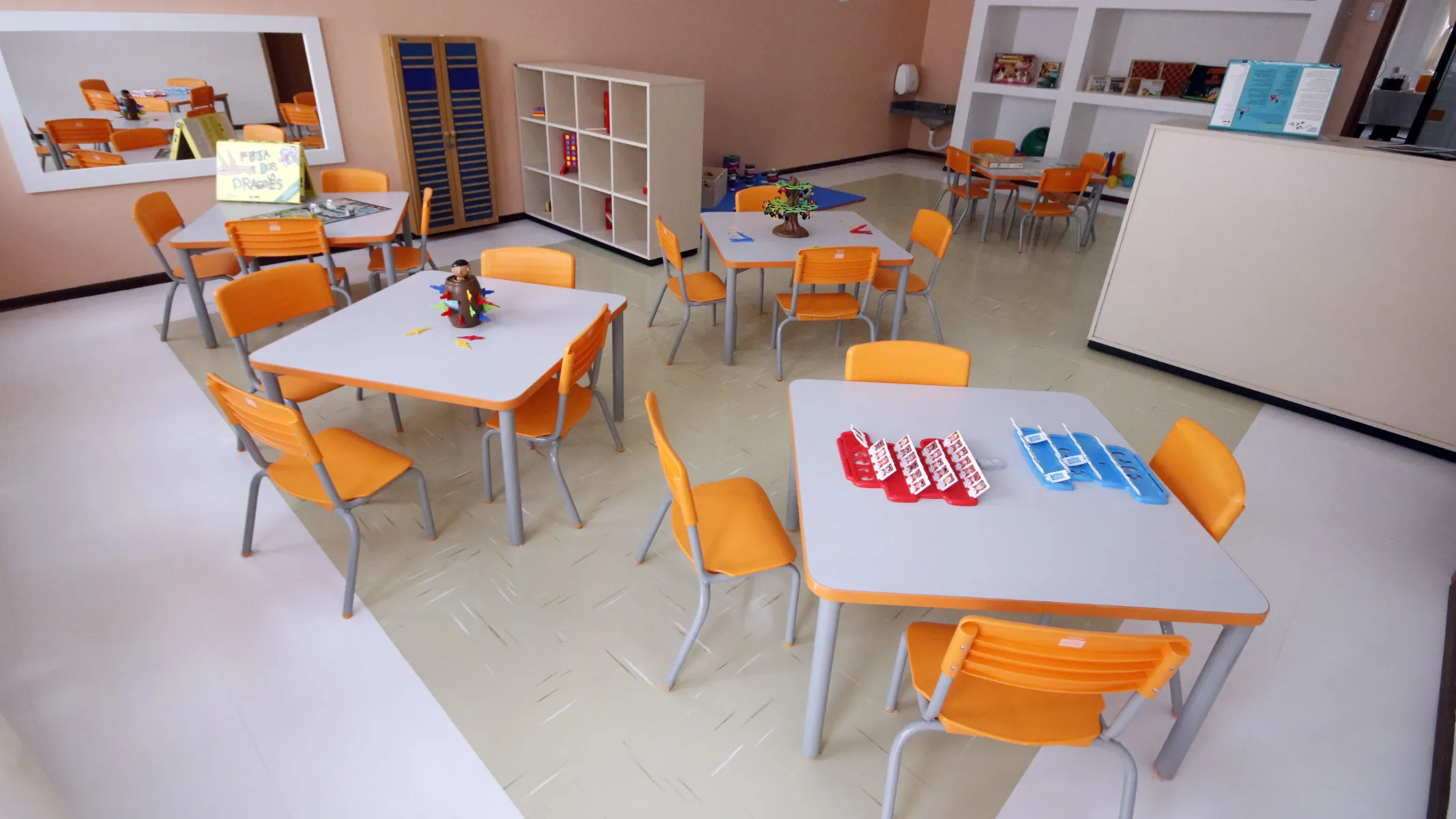 Fotografia em plano geral de uma sala de aula de educação infantil ou centro municipal de educação infantil (CMEI) organizada com mesinhas e cadeiras coloridas. Em primeiro plano e à direita, uma mesa quadrada com tampo cinza e pés metálicos é cercada por cadeiras laranjas, exibindo sobre ela jogos educativos ou materiais pedagógicos organizados. À esquerda e ao centro, outras mesas semelhantes com cadeiras laranjas estão dispostas pelo espaço, adornadas com pequenos brinquedos e enfeites pedagógicos. No plano intermediário, o ambiente amplo e bem iluminado conta com estantes organizadoras de madeira e nichos nas paredes com livros e materiais didáticos, além de um grande espelho emoldurado na parede à esquerda. Ao fundo, o piso claro e limpo e as paredes em tons neutros completam a ambientação interna da sala voltada para o atendimento infantil.