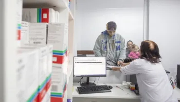 Fotografia em plano médio de um atendimento em um balcão de farmácia ou posto de saúde, com foco na interação entre uma profissional e um paciente. Em primeiro plano e à esquerda, prateleiras brancas repletas de caixas de medicamentos empilhadas ocupam a lateral da composição, apresentando rótulos coloridos. No plano intermediário, à direita, uma atendente vestindo jaleco branco está de costas para a câmera, entregando documentos ou receitas através de uma abertura envidraçada. Do outro lado do balcão, um jovem com capuz cinza e gorro atende ao chamado, enquanto uma mulher e uma criança aguardam logo atrás. Na bancada à frente da atendente, há um computador ligado, teclado e materiais de atendimento. Ao fundo, paredes claras e a iluminação interna completam o ambiente institucional de saúde sob luz difusa.