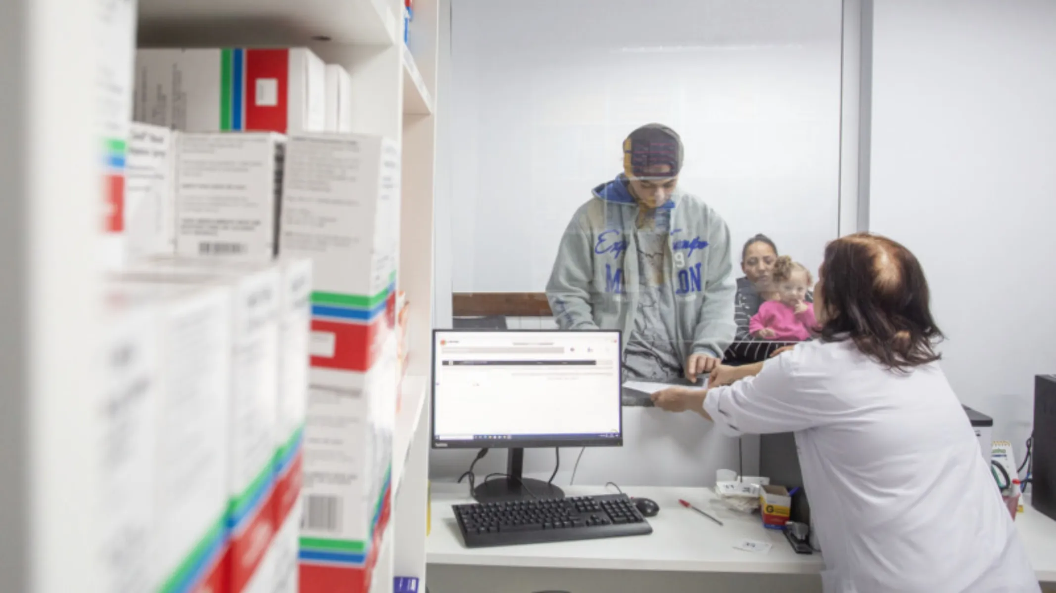 Fotografia em plano médio de um atendimento em um balcão de farmácia ou posto de saúde, com foco na interação entre uma profissional e um paciente. Em primeiro plano e à esquerda, prateleiras brancas repletas de caixas de medicamentos empilhadas ocupam a lateral da composição, apresentando rótulos coloridos. No plano intermediário, à direita, uma atendente vestindo jaleco branco está de costas para a câmera, entregando documentos ou receitas através de uma abertura envidraçada. Do outro lado do balcão, um jovem com capuz cinza e gorro atende ao chamado, enquanto uma mulher e uma criança aguardam logo atrás. Na bancada à frente da atendente, há um computador ligado, teclado e materiais de atendimento. Ao fundo, paredes claras e a iluminação interna completam o ambiente institucional de saúde sob luz difusa.