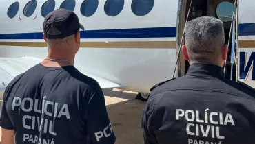 Fotografia em plano médio de dois policiais civis de costas, posicionados em frente a uma aeronave oficial da corporação. Em primeiro plano e à esquerda, um policial de costas veste camiseta preta com inscrições em destaque nas costas e na manga com os dizeres "POLÍCIA CIVIL" e "PARANÁ", além de boné escuro na cabeça. À direita, outro policial de costas veste uma camisa operacional preta com a mesma identificação institucional "POLÍCIA CIVIL PARANÁ" estampada nas costas em letras brancas. No plano intermediário, ambos estão diante de uma aeronave de pequeno porte de pintura branca, com detalhes em azul e dourado na fuselagem, janelas ovais e a porta aberta com escada de acesso posicionada. Ao fundo, o pátio aeroportuário sob luz solar diurna completa a cena da operação policial.