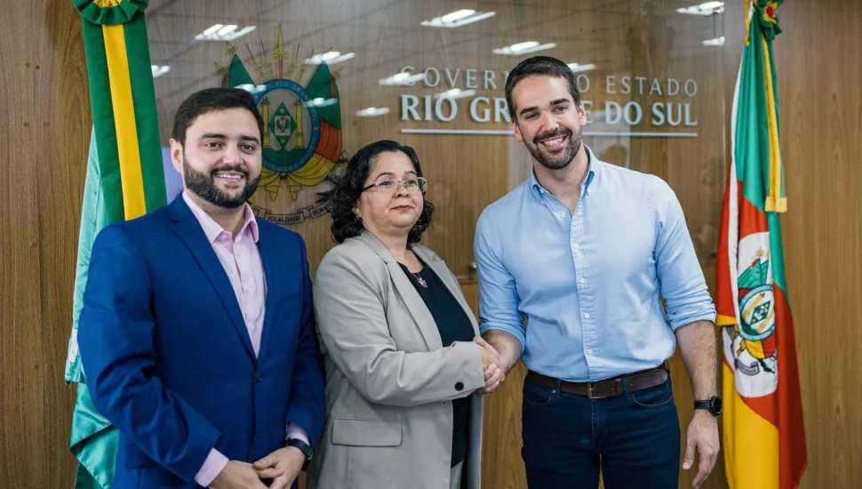 Fotografia em plano médio de três pessoas em um ambiente institucional, reunidas para um aperto de mãos diante de divisórias de vidro e bandeiras oficiais.Em primeiro plano e à esquerda, um homem com barba e cabelo escuros veste terno azul-marinho e camisa social rosa clara, mantendo os braços cruzados à frente. Ao centro, uma mulher de óculos, cabelos escuros e blazer cinza aperta a mão do homem à direita. À direita, o governador Eduardo Leite sorri para a câmera, vestindo camisa social azul-clara de mangas dobradas, calça jeans escura e cinto marrom. No plano intermediário, o encontro ocorre em uma sala institucional decorada com paredes revestidas em madeira e um painel de vidro ao fundo que exibe a inscrição em letras prateadas "GOVERNO DO ESTADO RIO GRANDE DO SUL" e o brasão oficial do estado aplicado em destaque. Ao fundo, bandeiras oficiais do Rio Grande do Sul, com suas faixas verdes, vermelhas e amarelas, flanqueiam as laterais da cena sob iluminação interna difusa.