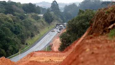Fotografia em plano geral de uma rodovia asfaltada cercada por vegetação densa e áreas de terra vermelha exposta, onde ocorreu um acidente de trânsito.Em primeiro plano e à direita, encostas de terra vermelha revolvida e vegetação rasteira ocupam a margem da pista, indicando obras ou movimentação de solo no local. No plano intermediário, a rodovia de pista dupla exibe traçado sinuoso com sinalização horizontal amarela. Ao fundo, na pista, veem-se veículos parados e caminhões envolvidos em um acidente, cercados por cones de sinalização e equipes de atendimento. Ao fundo, colinas cobertas por florestas e árvores frondosas estendem-se até o horizonte sob um dia nublado.
