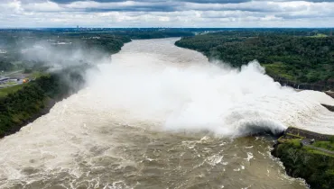 Fotografia aérea panorâmica de grande escala retratando o vertedouro de uma grande usina hidrelétrica, com imensos volumes de água espumosa sendo liberados no leito do rio. Em primeiro plano, as águas caudalosas e barrentas do rio agitam-se fortemente, formando corredeiras e espuma densa à medida que avançam. No plano intermediário, à direita, a estrutura de concreto da barragem descarrega uma enorme tromba d'água branca e vaporizada, criando uma imensa nuvem de gotículas e névoa que se espalha sobre o canal. Margens verdes e arborizadas delimitam os dois lados do curso d'água. Ao fundo, o rio segue seu leito em direção ao horizonte sob um céu nublado com nuvens densas e acinzentadas.