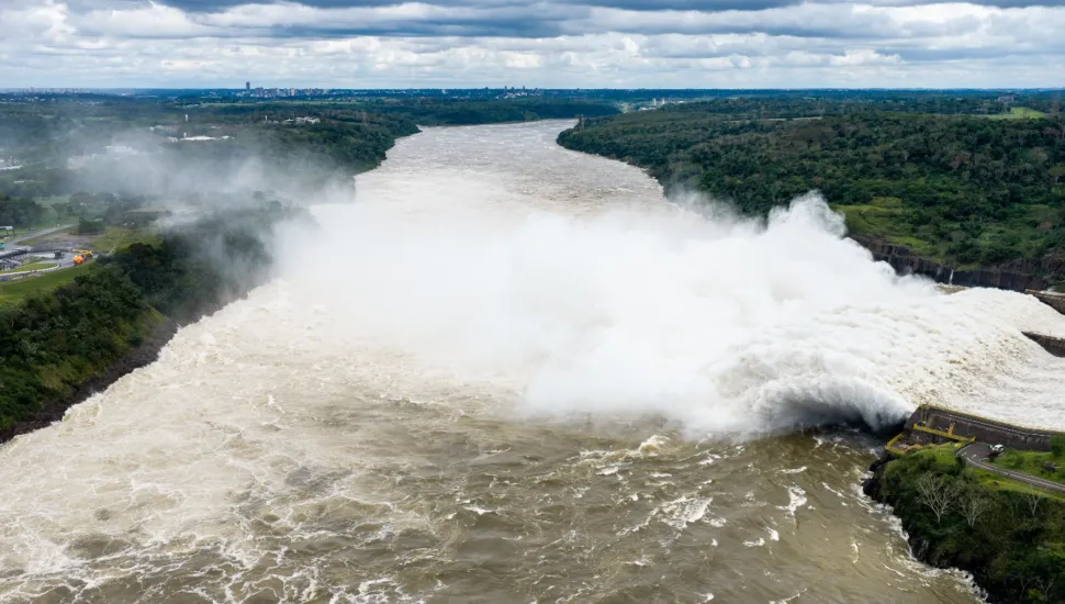 Fotografia aérea panorâmica de grande escala retratando o vertedouro de uma grande usina hidrelétrica, com imensos volumes de água espumosa sendo liberados no leito do rio. Em primeiro plano, as águas caudalosas e barrentas do rio agitam-se fortemente, formando corredeiras e espuma densa à medida que avançam. No plano intermediário, à direita, a estrutura de concreto da barragem descarrega uma enorme tromba d'água branca e vaporizada, criando uma imensa nuvem de gotículas e névoa que se espalha sobre o canal. Margens verdes e arborizadas delimitam os dois lados do curso d'água. Ao fundo, o rio segue seu leito em direção ao horizonte sob um céu nublado com nuvens densas e acinzentadas.