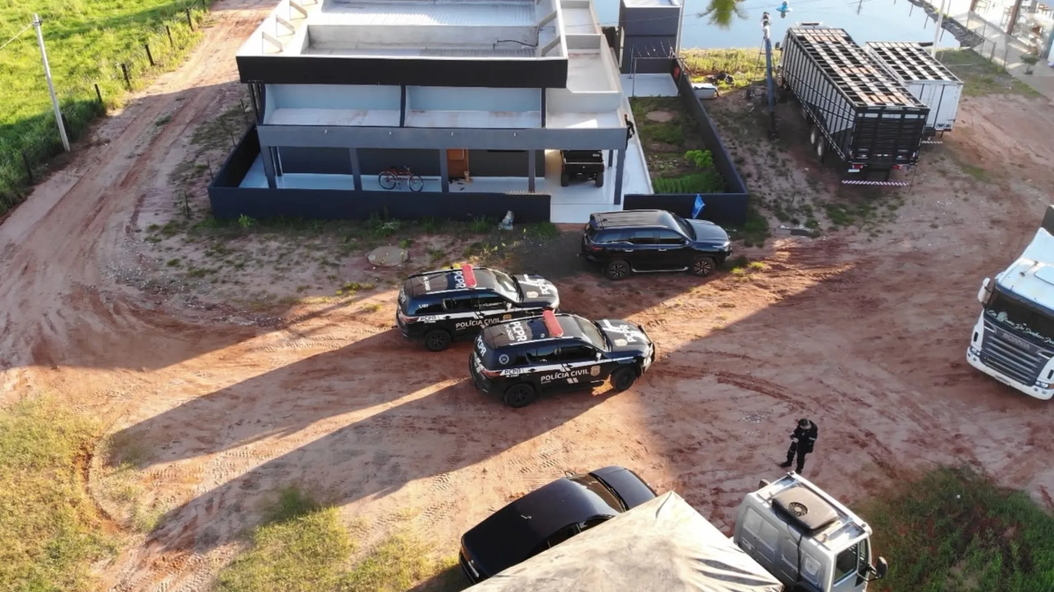Fotografia aérea em plano geral de uma operação policial em uma área residencial com terreno de terra batida, envolvendo viaturas oficiais e uma residência moderna. Em primeiro plano e na parte inferior, viaturas pretas caracterizadas da Polícia Civil estão estacionadas sobre o terreno de solo avermelhado, acompanhadas por um policial fardado em pé próximo a um caminhão. No plano intermediário, uma residência térrea de arquitetura contemporânea, com fachada em tons de cinza escuro e teto plano, destaca-se cercada por muros baixos e áreas verdes. Ao lado da casa e espalhados pelo terreno encontram-se mais automóveis, caminhões de grande porte e carrocerias estacionados. Ao fundo, a paisagem exibe áreas gramadas e margens de corpos d'água sob luz solar diurna clara.