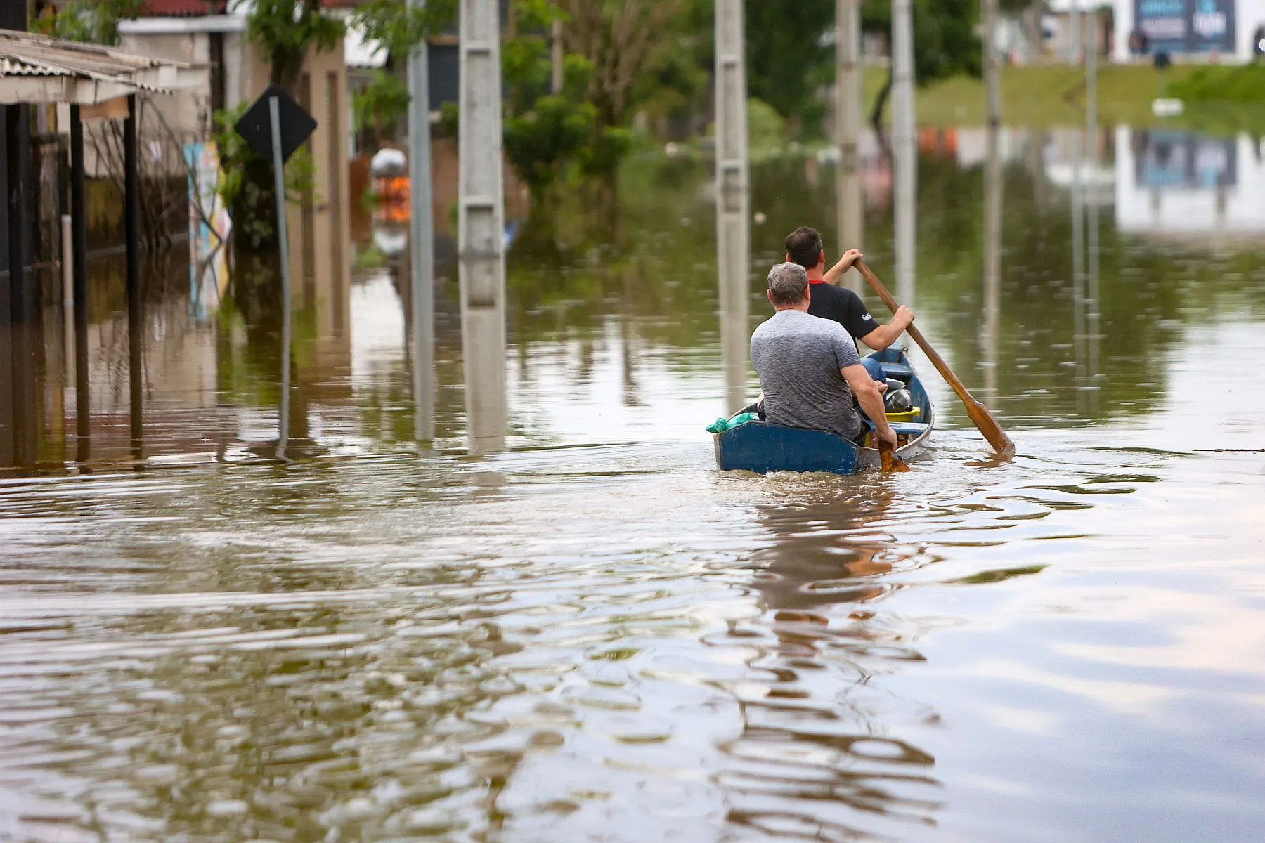 Imagem mostra uma rua alagada e dois homens remando numa canoa durante a enchente
