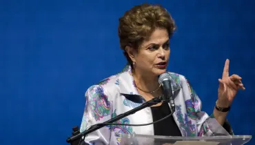 Fotografia em plano médio de Dilma Rousseff posicionada atrás de um púlpito durante um pronunciamento público. Em primeiro plano, Dilma Rousseff aparece com expressão séria e concentrada, gesticulando com a mão esquerda erguida e o indicador apontado para cima. Ela veste uma jaqueta acetinada de fundo claro estampada com padrões coloridos em tons de azul, verde e roxo, sobre uma blusa preta, além de um colar discreto e brincos pequenos. À sua frente, microfones fixados a um suporte metálico estão posicionados sobre a bancada transparente do púlpito. No plano intermediário, a figura da oradora destaca-se com nitidez sob iluminação frontal direcionada. Ao fundo, um painel liso em tom azul escuro ocupa todo o campo visual, criando um fundo neutro e uniforme para a cena.