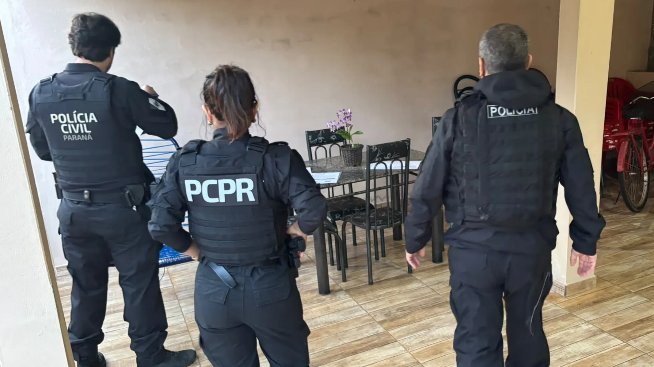 Fotografia em plano médio de três policiais civis de costas, posicionados em uma área coberta ou varanda residencial durante uma operação. Em primeiro plano, da esquerda para a direita: um policial à esquerda veste uniforme operacional preto com colete tático e as inscrições "POLÍCIA CIVIL PARANÁ" estampadas nas costas; ao centro, uma policial com os cabelos presos em rabo de cavalo veste uniforme preto e colete tático com a sigla "PCPR" em destaque; à direita, um policial veste jaqueta preta e colete tático com a identificação "POLICIAL" nas costas. Todos usam calças e botas pretas. No plano intermediário, o grupo encontra-se em um espaço com piso de cerâmica amadeirada, voltados para uma mesa de jantar com cadeiras pretas e um pequeno vaso com flores lilases sobre o tampo. Ao fundo, paredes em tons neutros e estruturas de uma residência completam o ambiente sob luz natural difusa.