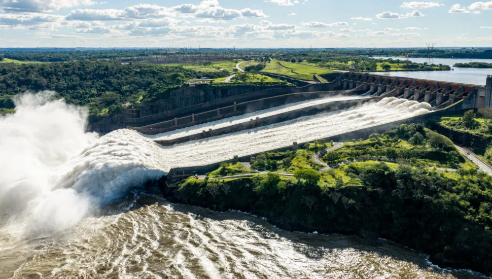 Fotografia aérea panorâmica em vista angular da estrutura do vertedouro de uma usina hidrelétrica de grande porte com as comportas abertas. Em primeiro plano e à esquerda, volumosos jatos de água branca e espumosa desaguam violentamente no rio, formando uma densa nuvem de vapor e névoa sobre a água barrenta agitada em tons de castanho e bege. No plano intermediário, as calhas de concreto inclinado do vertedouro direcionam o fluxo de água a partir da estrutura da barragem, situada à direita. Áreas verdes arborizadas e encostas com vegetação delimitam as margens do complexo de engenharia. Ao fundo, a albufeira estende-se sob a paisagem de relevo suave e áreas florestadas, sob um céu azul povoado por nuvens brancas e iluminação solar diurna.