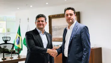 Fotografia em plano médio de um aperto de mãos institucional entre Sergio Moro e Ratinho Junior em um gabinete governamental. Em primeiro plano e à esquerda, Sergio Moro sorri para a câmara enquanto veste um terno preto e camisa social azul-clara aberta no colarinho. À direita, o governador Ratinho Junior veste um fato azul-marinho sobre camisa branca sem gravata, mantendo o olhar direcionado para a frente. Ambos cumprem um aperto de mãos no centro da composição. No plano intermediário, à esquerda, observa-se uma mesa de madeira com pequenos enfeites decorativos e, logo atrás, a bandeira do Brasil e a bandeira do Estado do Paraná dispostas verticalmente em mastros. Ao fundo, uma parede clara exibe um quadro com moldura dourada e entalhada, sob iluminação embutida de teto.