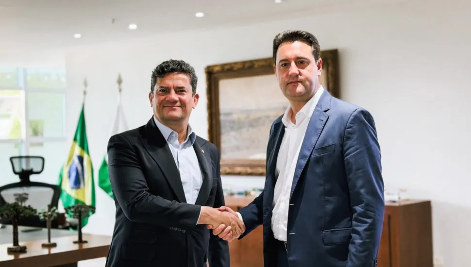 Fotografia em plano médio de um aperto de mãos institucional entre Sergio Moro e Ratinho Junior em um gabinete governamental. Em primeiro plano e à esquerda, Sergio Moro sorri para a câmara enquanto veste um terno preto e camisa social azul-clara aberta no colarinho. À direita, o governador Ratinho Junior veste um fato azul-marinho sobre camisa branca sem gravata, mantendo o olhar direcionado para a frente. Ambos cumprem um aperto de mãos no centro da composição. No plano intermediário, à esquerda, observa-se uma mesa de madeira com pequenos enfeites decorativos e, logo atrás, a bandeira do Brasil e a bandeira do Estado do Paraná dispostas verticalmente em mastros. Ao fundo, uma parede clara exibe um quadro com moldura dourada e entalhada, sob iluminação embutida de teto.