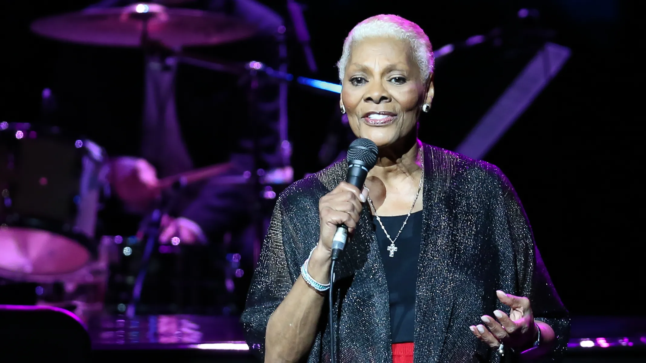 Fotografia em plano médio da cantora Dionne Warwick a atuar num palco, a segurar um microfone durante uma apresentação musical ao vivo. Em primeiro plano e ao centro, Dionne Warwick aparece com cabelos curtos e grisalhos, maquilhagem discreta, brincos pequenos e um colar com pendente em forma de cruz. Ela veste uma sobreposição brilhante e plissada de tom escuro sobre blusa preta, segurando o microfone com a mão direita junto à boca enquanto gesticula suavemente com a mão esquerda aberta. No plano intermediário, a artista é focada sob iluminação cénica direcionada, sobressaindo da penumbra do palco. Ao fundo, na penumbra com luzes em tons de violeta e azul, veem-se desfocados os elementos de uma bateria musical, incluindo pratos e tambores, além de parte da estrutura da banda de suporte.