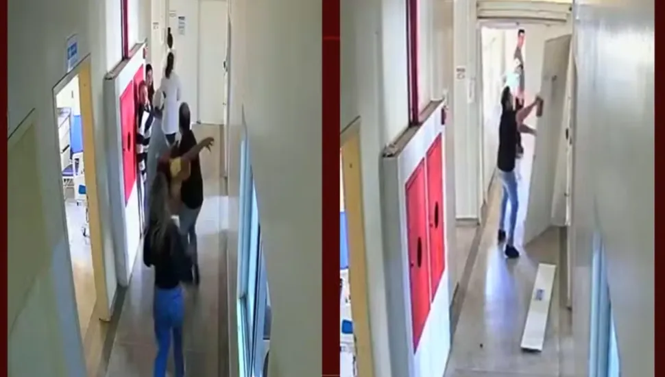 Fotografia composta por dois quadros de câmeras de segurança (CCTV) posicionados lado a lado, registrando uma altercação física no corredor de uma unidade de saúde ou hospital. Em primeiro plano, no quadro da esquerda, um corredor claro exibe portas laterais e armários vermelhos de emergência fixados na parede. Pessoas movimentam-se pelo espaço, destacando-se uma mulher de costas de calça jeans azul e blusa preta, enquanto homens ao centro aparecem em meio a uma discussão ou confronto corporal.No quadro da direita, a partir de um ângulo diferente do mesmo corredor, um homem de camiseta preta e calça jeans ergue os braços contra uma divisória ou porta branca, em atitude hostil ou de confronto. Outras pessoas observam a cena ao fundo da passagem.Ao fundo, em ambos os quadros, o corredor estende-se com paredes claras sob iluminação artificial de lâmpadas fluorescentes de teto.