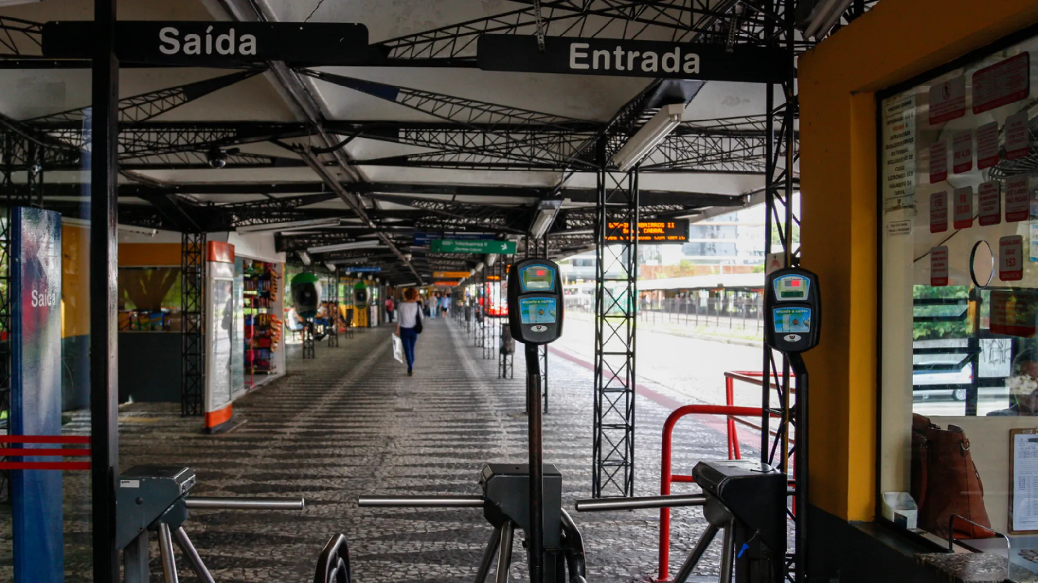 Fotografia em plano geral do interior de uma estação-tubo de transporte coletivo em Curitiba, captada a partir da catraca de acesso. Em primeiro plano, duas catracas metálicas com leitores eletrônicos pretos e visores iluminados marcam a entrada da estação. Acima delas, uma placa sinaliza claramente as direções com os termos "Saída" à esquerda e "Entrada" à direita. À direita, parte da cabine amarela de atendimento e bilheteria exibe informativos fixados no vidro. No plano intermediário, o interior da estação estende-se em formato tubular com piso de paralelepípedos e estruturas metálicas arqueadas no teto. Uma pessoa caminha ao fundo em direção oposta, enquanto balcões laterais e painéis informativos acompanham a extensão da plataforma. Ao fundo, a abertura transparente da estação exibe a claridade do dia e a paisagem urbana externa sob iluminação natural.