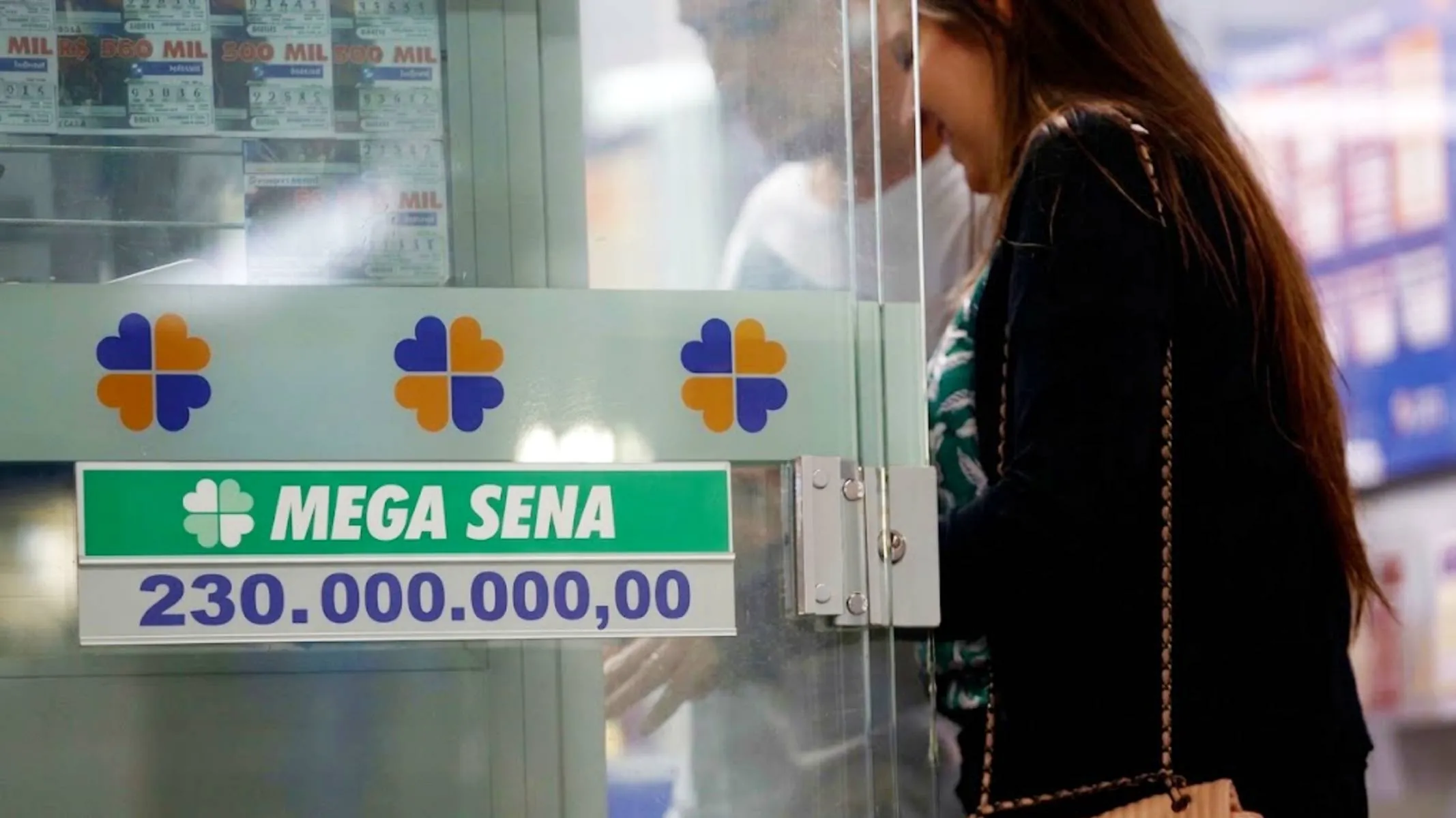 Imagem mostra uma lotérica com uma mulher e um homem apostando. Eles estão atrás de uma placa do prêmio de R$ 230 milhões.