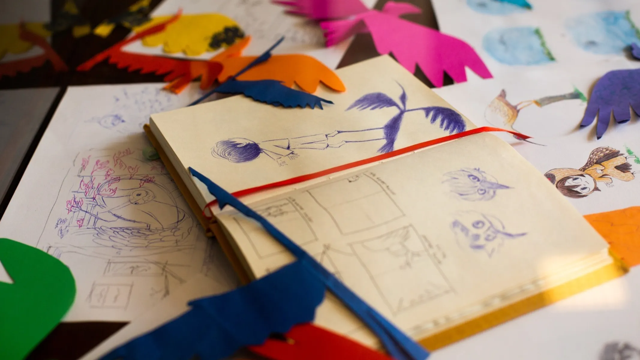 Fotografia em plano aproximado (close-up) e ângulo superior de uma mesa de trabalho repleta de esboços artísticos, recortes de papel colorido e cadernos de desenho abertos. Em primeiro plano e ao centro, um caderno de capa amarela está aberto, exibindo páginas de papel creme com esboços a caneta azul ou grafite, incluindo estudos de personagens, rascunhos de histórias em quadrinhos (storyboard) e recortes de papel azul recortados em formato de silhuetas de aves que cruzam o papel. Um marcador de página em fita vermelha estende-se sobre as folhas. No plano intermediário, espalham-se folhas de papel avulsas com desenhos coloridos, ilustrações de figuras humanas, aves e cenários, além de diversos recortes artesanais de papel em cores vivas como rosa, laranja, amarelo e verde. Ao fundo, a superfície escura da mesa serve de base para a composição iluminada por luz natural suave.