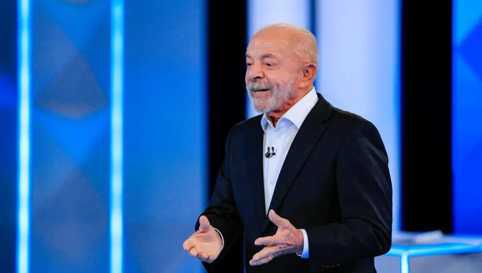 Fotografia em plano médio de Luiz Inácio Lula da Silva em pé, gesticulando enquanto discursa em um estúdio televisivo ou auditório com cenário iluminado em tons de azul.Em primeiro plano e ao centro, Lula aparece sorrindo levemente com o olhar voltado para a esquerda. Ele veste um fato escuro, camisa social branca com os primeiros botões abertos e um pequeno microfone de lapela preso ao colarinho. Suas mãos estão abertas à altura da cintura, em gesto expressivo de fala. No plano intermediário, o retratado destaca-se sob forte iluminação frontal de estúdio, focada em sua figura.Ao fundo, painéis verticais com iluminação em LED azul e faixas verticais claras compõem a cenografia do ambiente sob luz difusa.