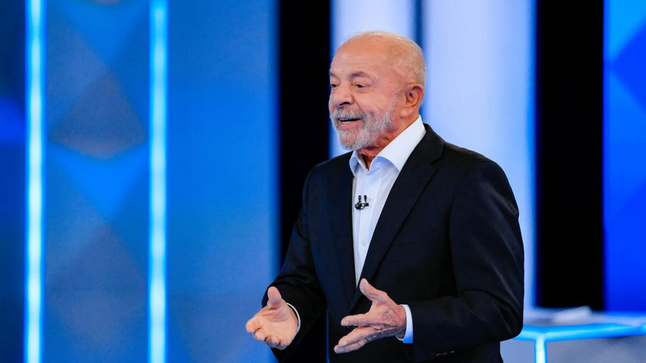 Fotografia em plano médio de Luiz Inácio Lula da Silva em pé, gesticulando enquanto discursa em um estúdio televisivo ou auditório com cenário iluminado em tons de azul.Em primeiro plano e ao centro, Lula aparece sorrindo levemente com o olhar voltado para a esquerda. Ele veste um fato escuro, camisa social branca com os primeiros botões abertos e um pequeno microfone de lapela preso ao colarinho. Suas mãos estão abertas à altura da cintura, em gesto expressivo de fala. No plano intermediário, o retratado destaca-se sob forte iluminação frontal de estúdio, focada em sua figura.Ao fundo, painéis verticais com iluminação em LED azul e faixas verticais claras compõem a cenografia do ambiente sob luz difusa.
