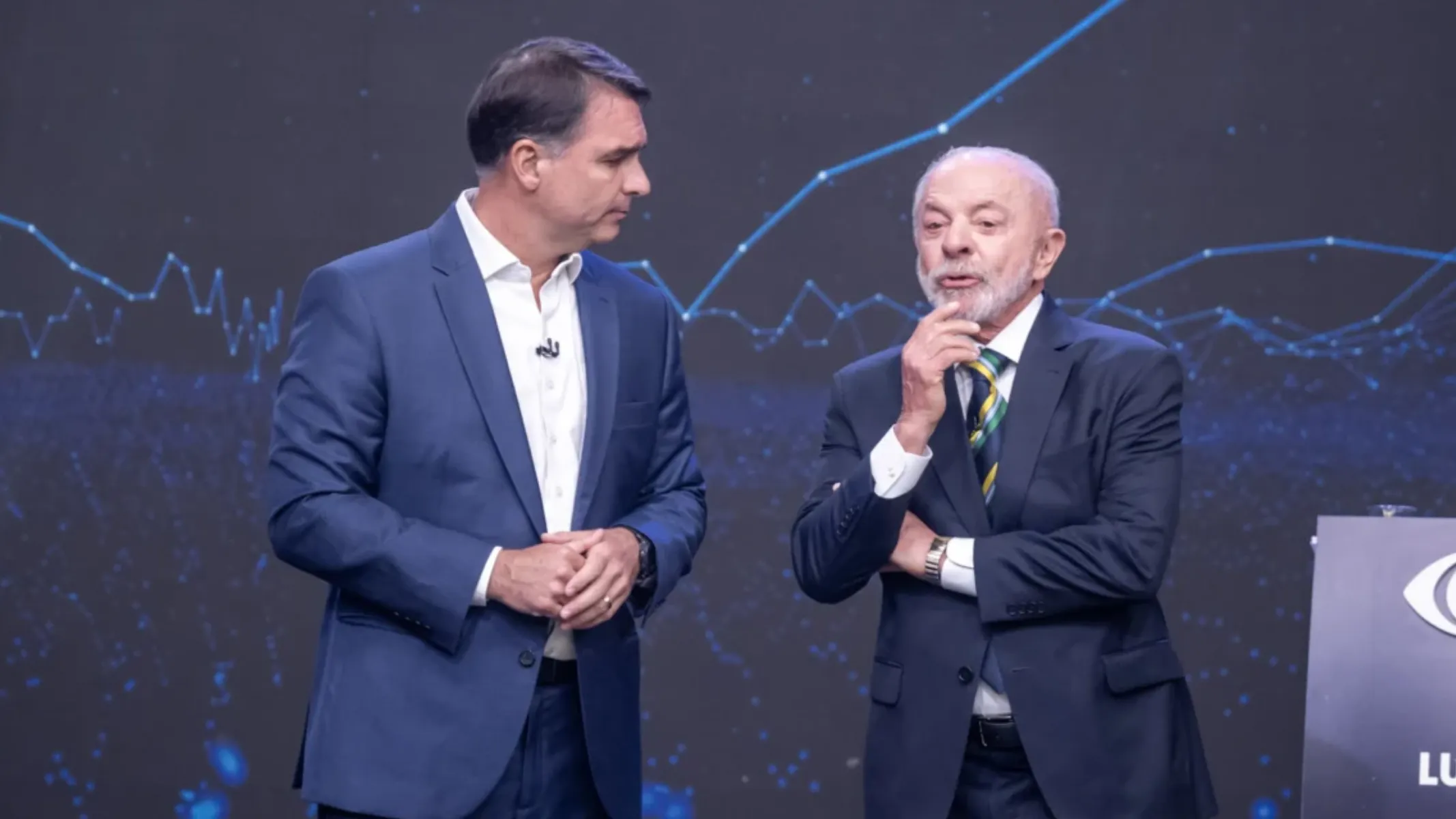 Fotografia em plano médio do senador Flávio Bolsonaro e do presidente Luiz Inácio Lula da Silva lado a lado durante um debate político televisivo. Em primeiro plano e à esquerda, Flávio Bolsonaro veste um fato azul-marinho e camisa social branca de colarinho aberto sem gravata, mantendo as mãos entrelaçadas à frente e o olhar direcionado para Lula. À direita, Lula veste um fato escuro, camisa branca e gravata com listras em verde e amarelo, com a mão direita elevada ao queixo em atitude reflexiva enquanto fala. No plano intermediário, os dois líderes políticos posicionam-se em pé no palco do estúdio, com parte de um púlpito escuro e o logotipo da emissora visíveis no canto inferior direito. Ao fundo, um painel LED escuro exibe gráficos em rede de linhas e pontos azuis brilhantes sob iluminação cénica direcionada.