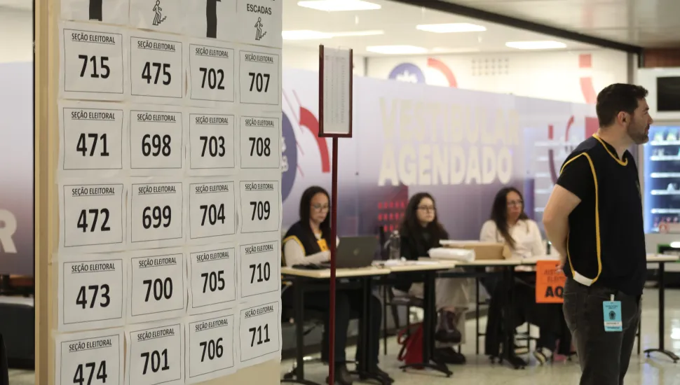 Fotografia em plano médio de uma seção de votação ou local de atendimento eleitoral, destacando um painel informativo com números de seções e funcionários organizados no espaço. Em primeiro plano e à esquerda, um grande painel vertical branco afixa vários papéis impressos com a indicação "SEÇÃO ELEITORAL" e numerações pretas correspondentes (como 715, 475, 702, 707, 471, entre outras) dispostas em grelha. Na extremidade direita do primeiro plano, um homem de perfil, vestindo uma camisola escura com debruns amarelos e calças jeans, observa o ambiente. Um crachá de identificação está preso à sua cintura. No plano intermediário, mesas de atendimento organizadas em fila acomodam três mulheres sentadas, que trabalham com computadores e documentos sob iluminação interna de escritório. Ao fundo, divisórias com painéis informativos exibem inscrições parciais, como a palavra "AGENDADO", compondo o cenário institucional do local de votação.