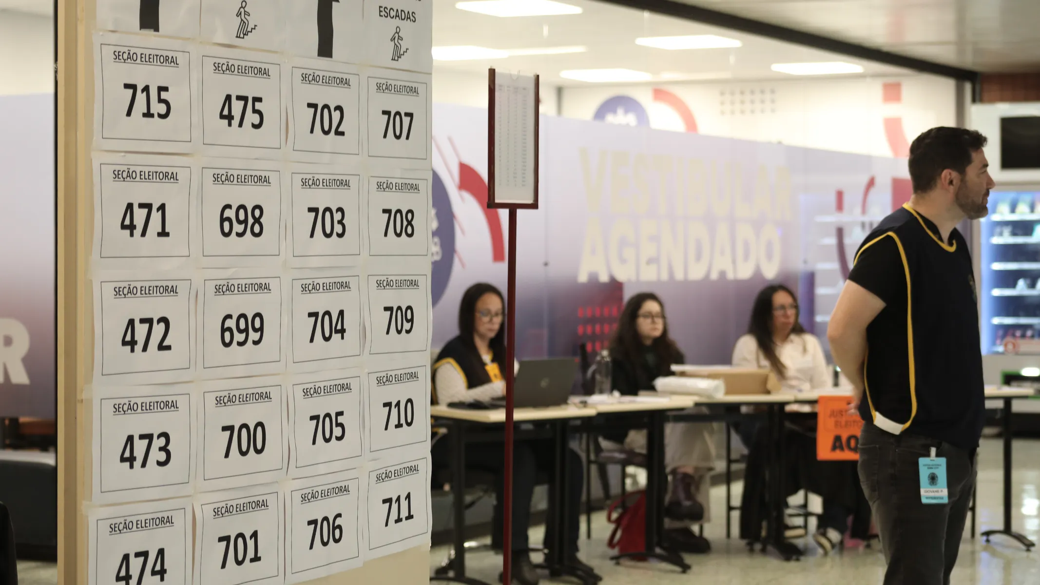 Fotografia em plano médio de uma seção de votação ou local de atendimento eleitoral, destacando um painel informativo com números de seções e funcionários organizados no espaço. Em primeiro plano e à esquerda, um grande painel vertical branco afixa vários papéis impressos com a indicação "SEÇÃO ELEITORAL" e numerações pretas correspondentes (como 715, 475, 702, 707, 471, entre outras) dispostas em grelha. Na extremidade direita do primeiro plano, um homem de perfil, vestindo uma camisola escura com debruns amarelos e calças jeans, observa o ambiente. Um crachá de identificação está preso à sua cintura. No plano intermediário, mesas de atendimento organizadas em fila acomodam três mulheres sentadas, que trabalham com computadores e documentos sob iluminação interna de escritório. Ao fundo, divisórias com painéis informativos exibem inscrições parciais, como a palavra "AGENDADO", compondo o cenário institucional do local de votação.