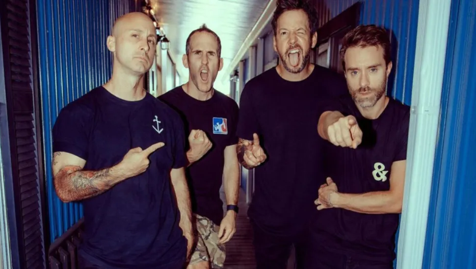 Fotografia em plano médio de uma banda de rock composta por quatro homens posando juntos num corredor estreito com paredes de metal azul. Em primeiro plano, à esquerda, um homem careca com tatuagens no braço veste uma t-shirt preta e aponta o dedo indicador para o próprio peito com expressão séria. Ao seu lado, o segundo homem, também de t-shirt preta, surge com a boca aberta numa expressão enérgica e o punho fechado. O terceiro homem, mais alto e igualmente de t-shirt preta, posa com a boca aberta e os olhos arregalados, fazendo uma careta divertida. À extrema direita, o quarto homem, com barba curta e t-shirt preta ostentando o símbolo de um e comercial (&), aponta diretamente o dedo indicador para a câmara com um olhar fixo. No plano intermediário, os quatro integrantes ocupam o corredor sob iluminação direta e lateral que destaca suas feições. Ao fundo, paredes verticais metálicas em tom de azul acinzentado delimitam o corredor do espaço exterior ou dos bastidores.