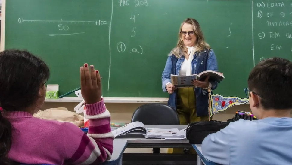 A imagem mostra um ambiente de sala de aula com professora e dois estudantes. Uma aluna está com a mão levantada.
