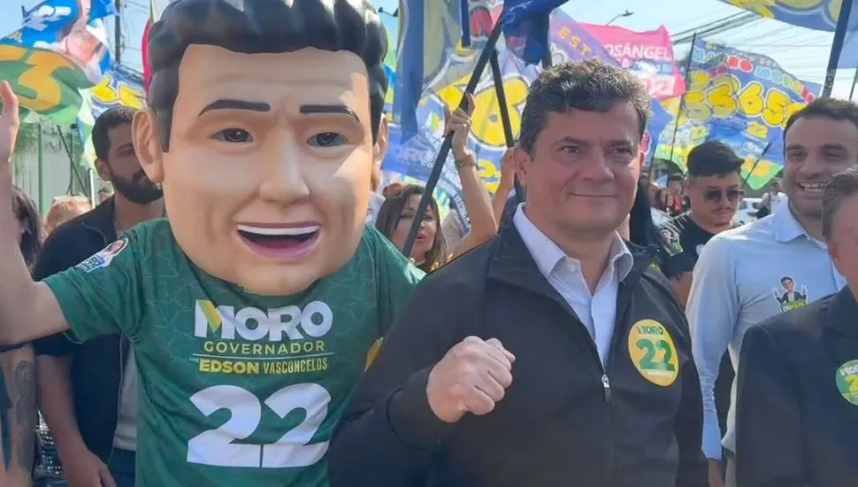 Sergio Moro caminha ao lado de apoiadores. No centro da imagem um boneco em tamanho real representa o senador, que está ao lado fazendo um gesto de força com as mãos, com o punho cerrrado