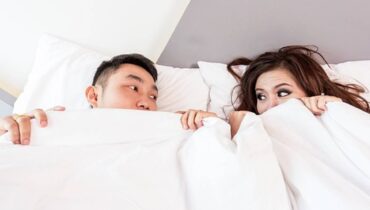 Casal na cama se cobre com uma coberta branca e troca olhares