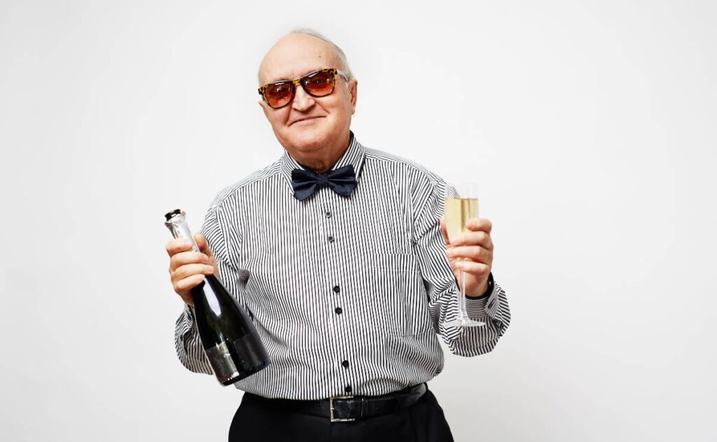 Imagem mostra um idoso rindo com uma champagne e uma taça nas mãos