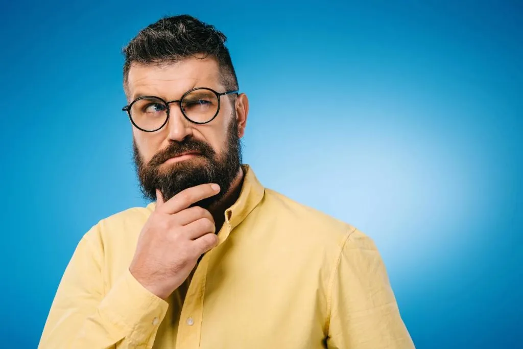 Imagem mostra um homem usando camisa amarela em um fundo azul. ele tem barba, usa óculos e está com a mão no queixo com cara de desconfiado.
