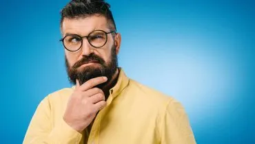 Imagem mostra um homem usando camisa amarela em um fundo azul. ele tem barba, usa óculos e está com a mão no queixo com cara de desconfiado.