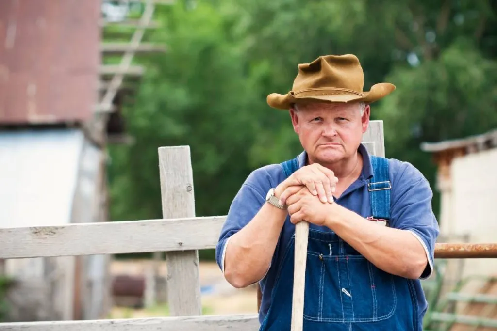 Imagem mostra um fazendeiro apoiado em uma enxada em uma fazenda.