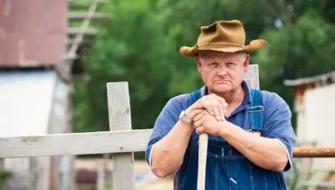 Imagem mostra um fazendeiro apoiado em uma enxada em uma fazenda.