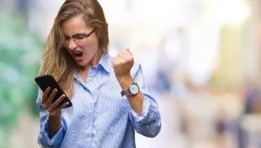 Imagem mostra uma mulher com raiva olhando para um celular. Ela está de camisa azul e com um relógio no pulso.