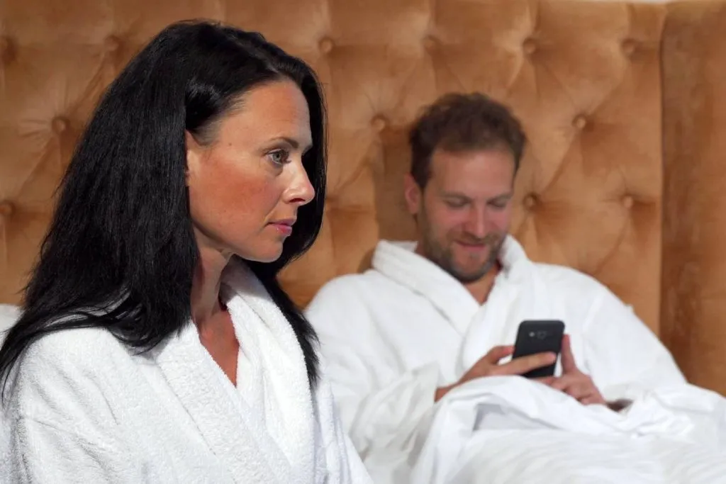Imagem mostra um casal em uma cama de hotel. Ele está no celular e ela brava. Ambos usam um roupão branco