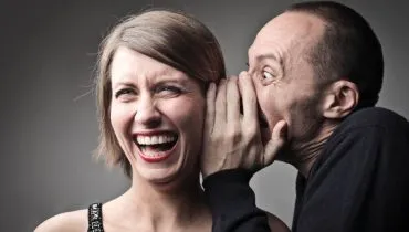 Imagem mostra um homem cochichando na orelha de uma mulher que está rindo.