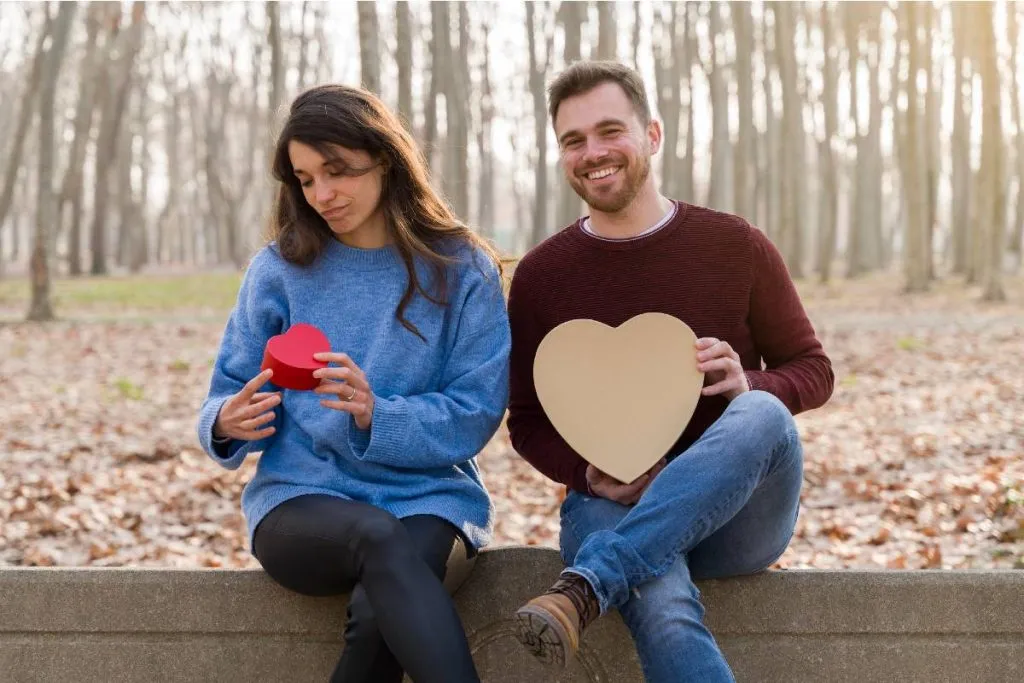Imagem mostra um casal de namorados trocando presentes em um parque. Ela está de camisa azul segurando um presentinho e ele de camisa bordô segurando um presente maior. Ela está decepcionada.