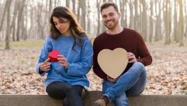 Imagem mostra um casal de namorados trocando presentes em um parque. Ela está de camisa azul segurando um presentinho e ele de camisa bordô segurando um presente maior. Ela está decepcionada.