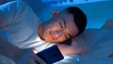 Imagem mostra um rapaz usando o celular a noite deitado na cama.