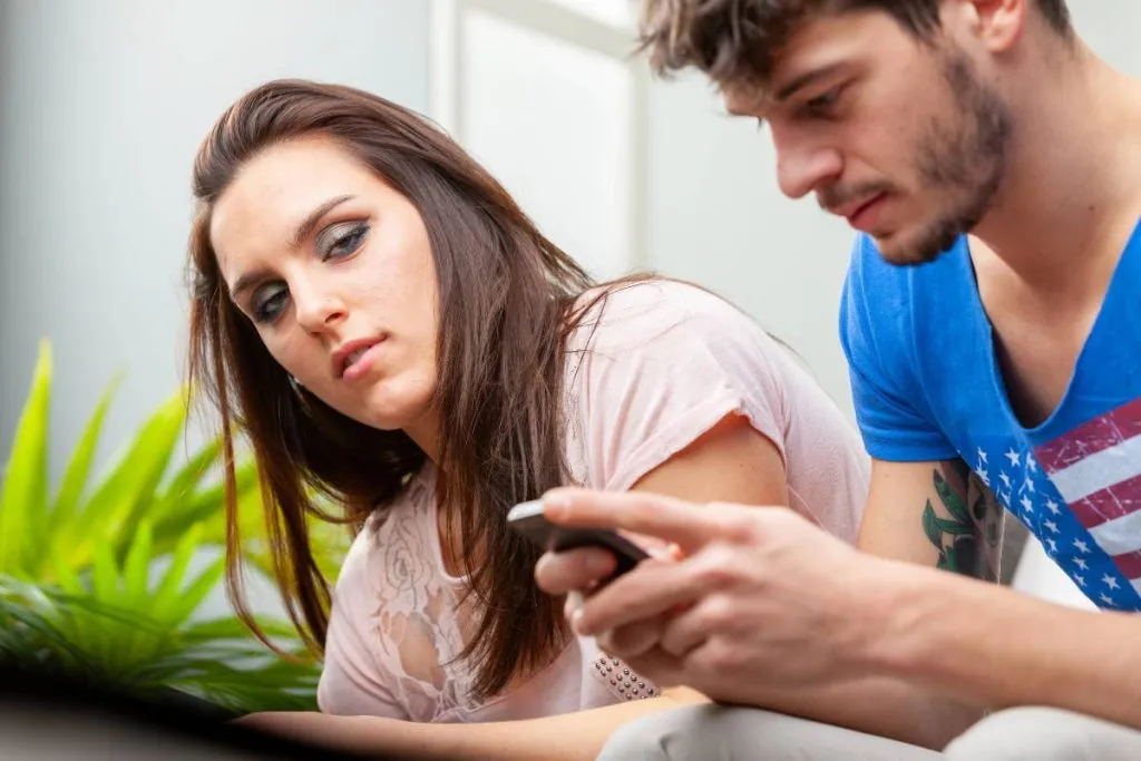 Imagem mostra um casal durante uma briga. Ele olha o celular e ela está com uma cara de desconfiada.
