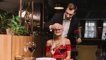 Imagem mostra um casal em um jantar romântico. Ele esta fazendo uma surpresa para ela, com uma flor na mão